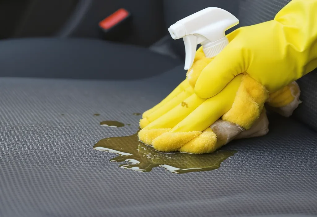 Close-up of a mechanic's hand using a microfiber cloth to clean a greasy stain from light gray car seat fabric.