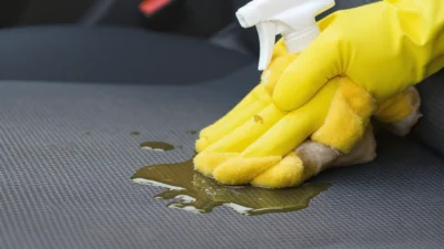 Close-up of a mechanic's hand using a microfiber cloth to clean a greasy stain from light gray car seat fabric.